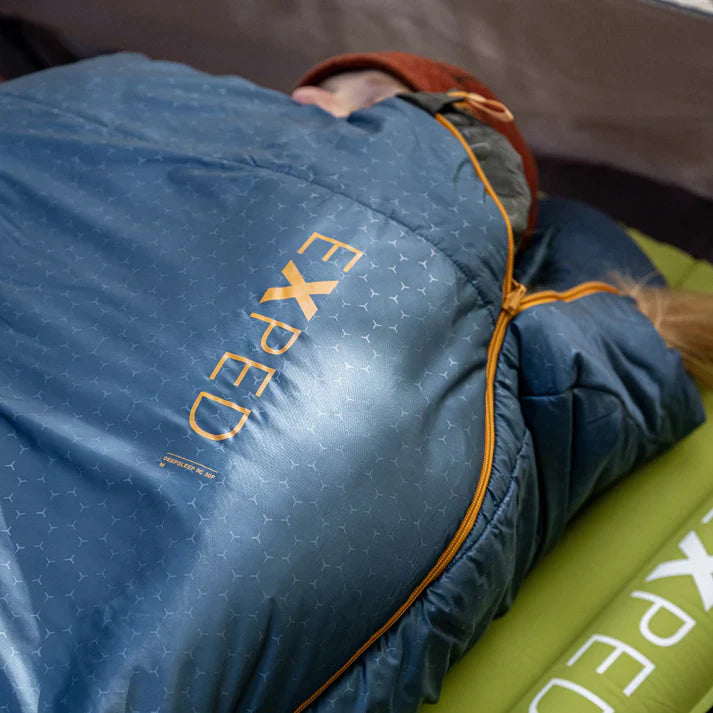 Person lying in a blue sleeping bag with 'Exped' branding on a green sleeping pad.
