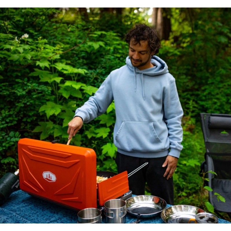 Person using an orange camping stove outdoors with greenery in the background
