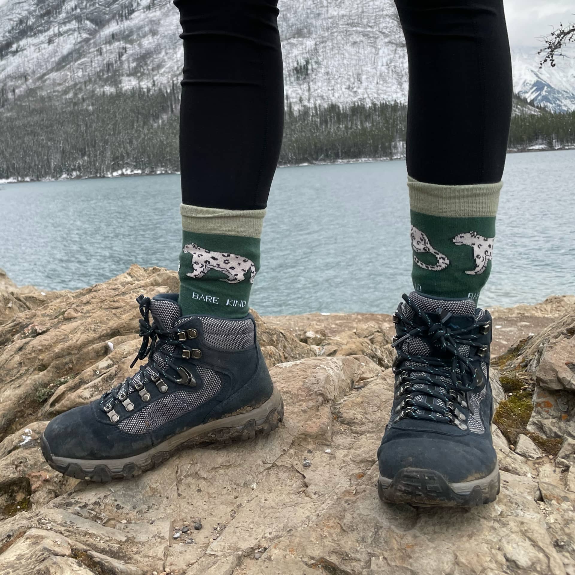 View of feet standing wearing walking boots and green Bare Kind socks with a snow leopard print.